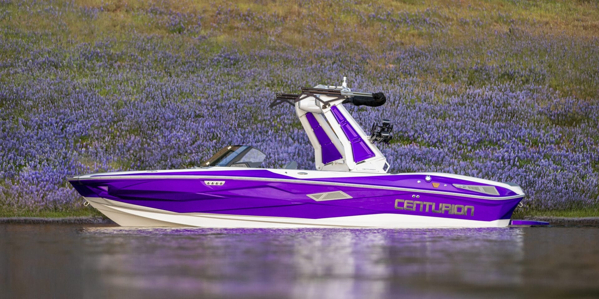 A purple and white Centurion speedboat is stationary on calm water, with a grassy, purple-flowered hillside in the background.