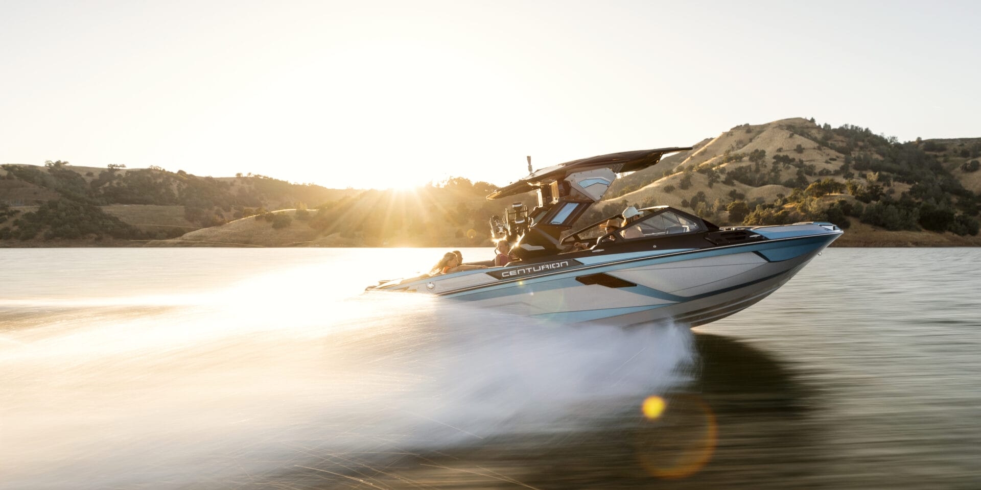 A speedboat moves quickly across a lake at sunset, with hills and trees visible in the background.