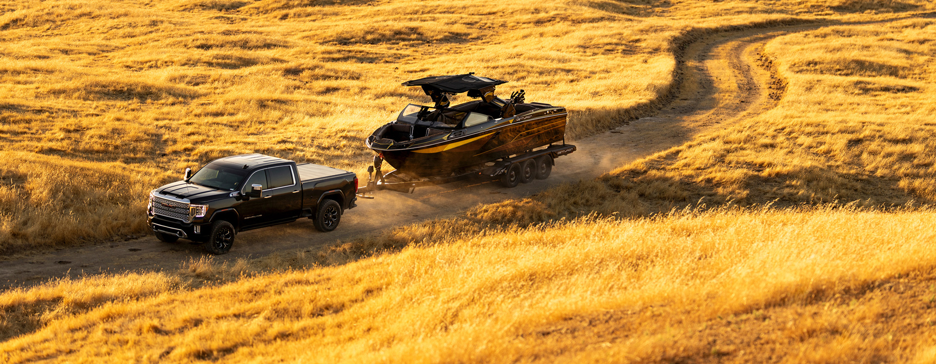 A black pickup truck tows a Centurion Ri230 Wake Boat on a trailer along a dirt road winding through a field of tall, yellow grass.