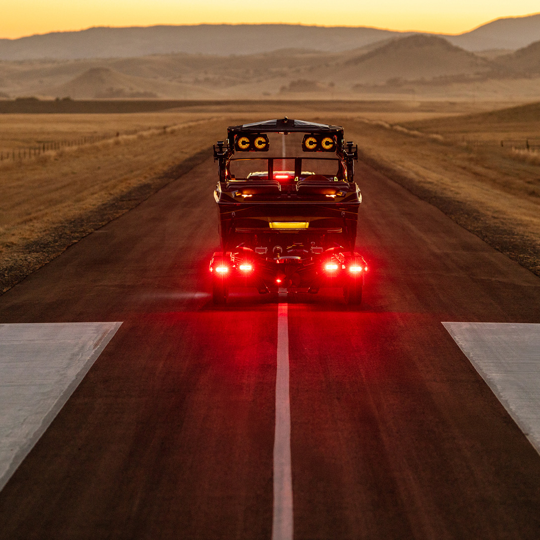 A Centurion Ri230 with bright red taillights travels down a rural road at sunset, surrounded by open fields and distant hills.