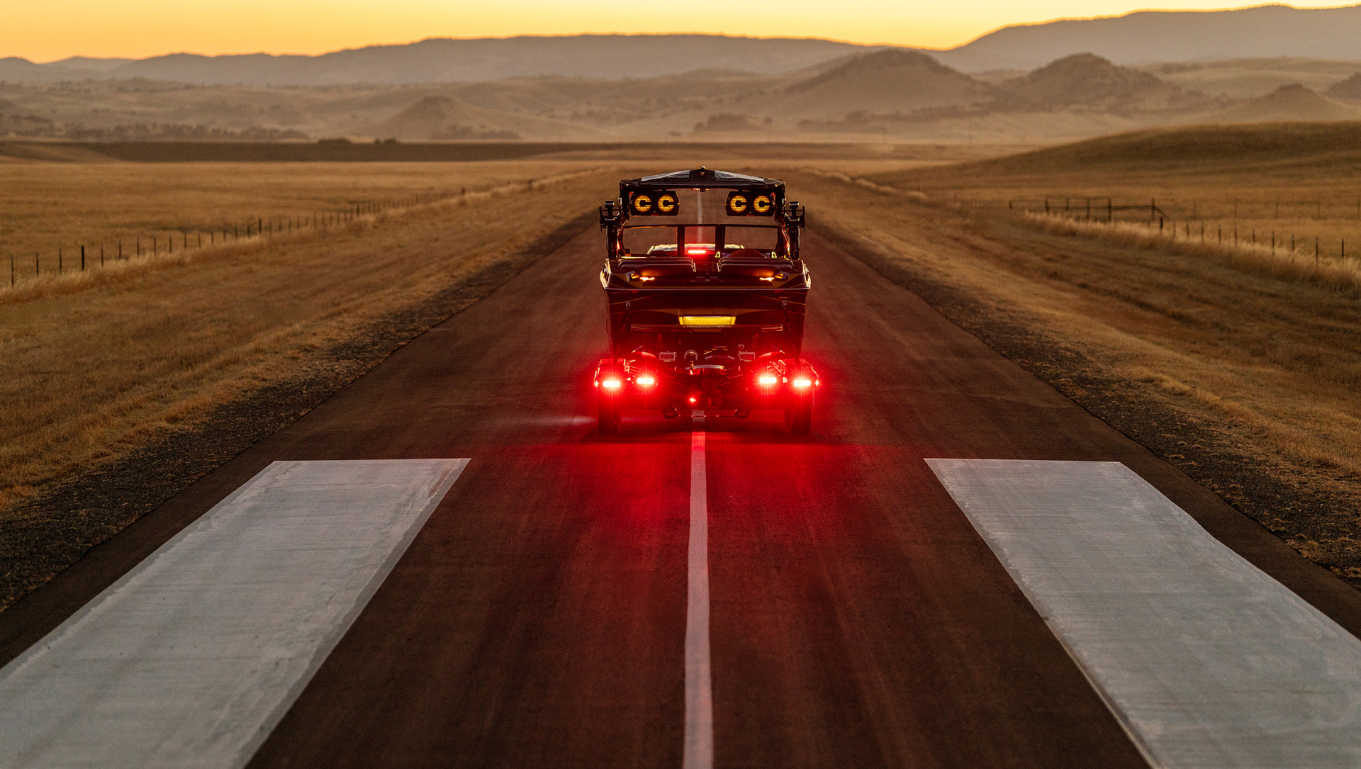 A Centurion Ri230 with bright red taillights travels down a rural road at sunset, surrounded by open fields and distant hills.