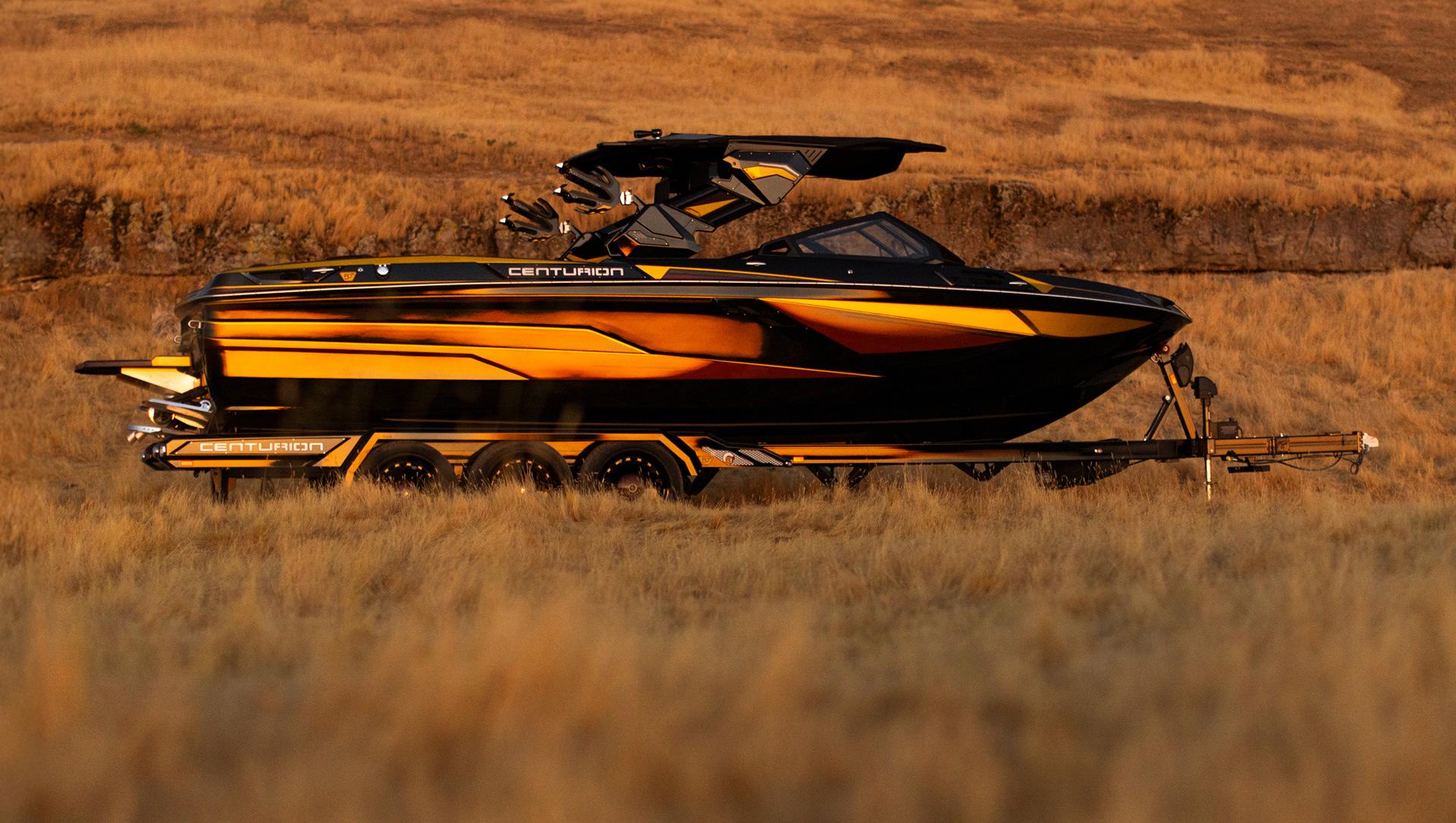 A black and yellow Centurion Ri230 Wake Boat on a trailer is parked in a dry grassy field, with hills in the background.