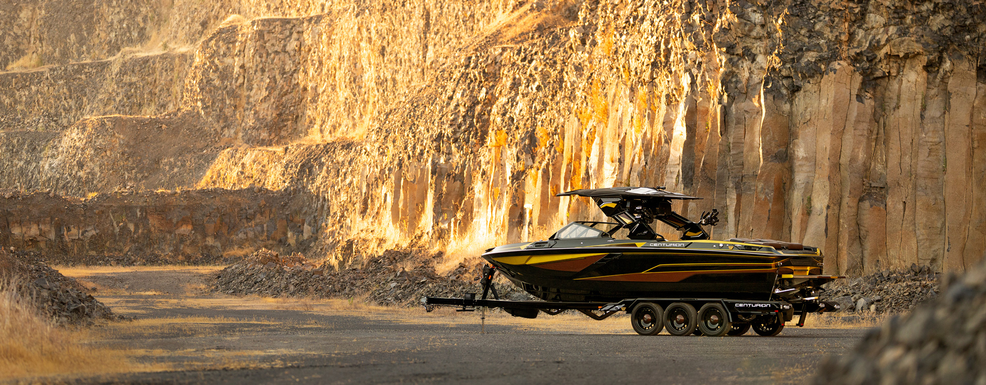A black and yellow Centurion Ri230 Wake Boat on a trailer is parked on a road beside tall, rocky cliffs in a sunlit, arid landscape.