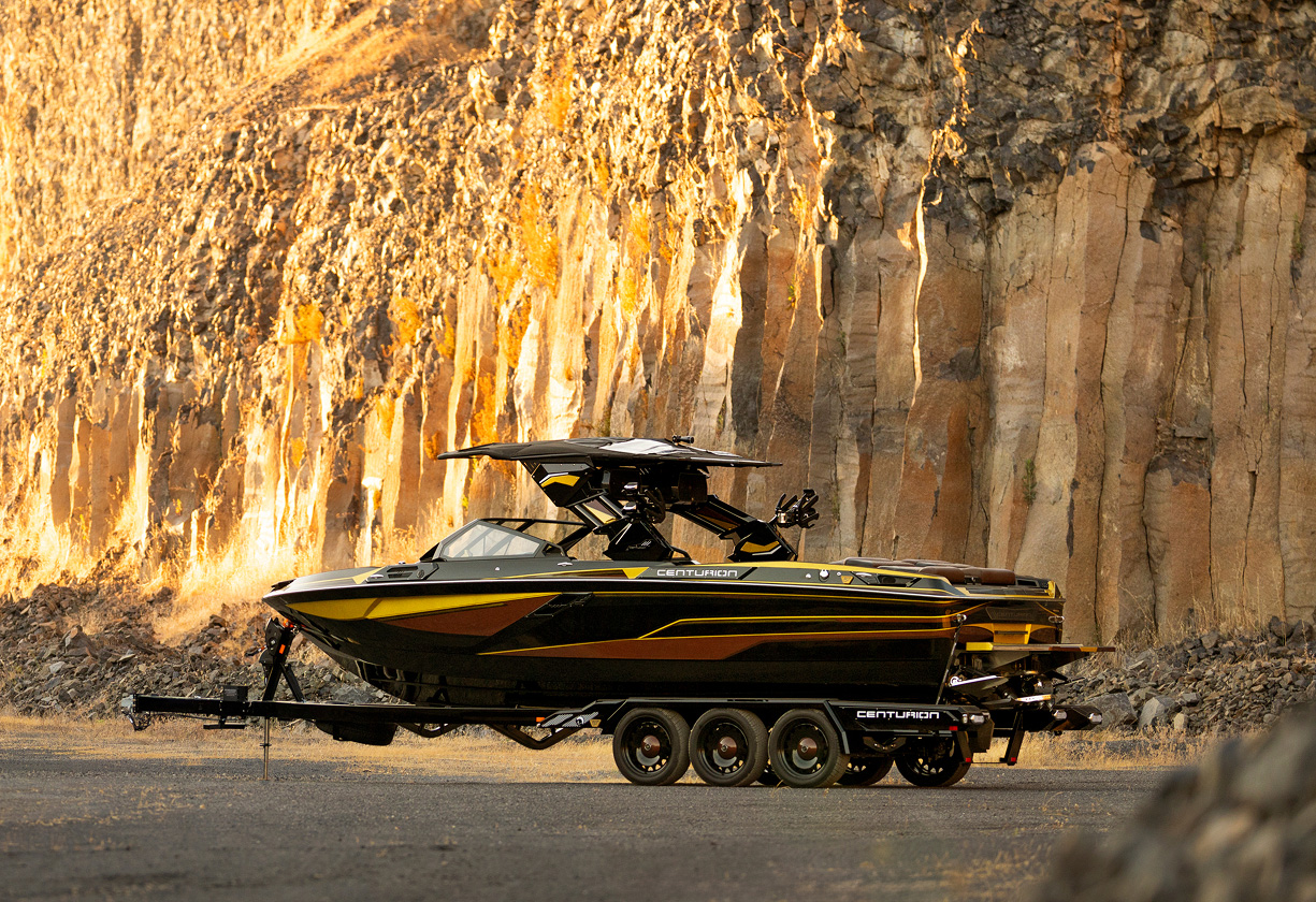 A black and yellow Centurion Ri230 Wake Boat is parked on a trailer beside rugged, sunlit rock cliffs.