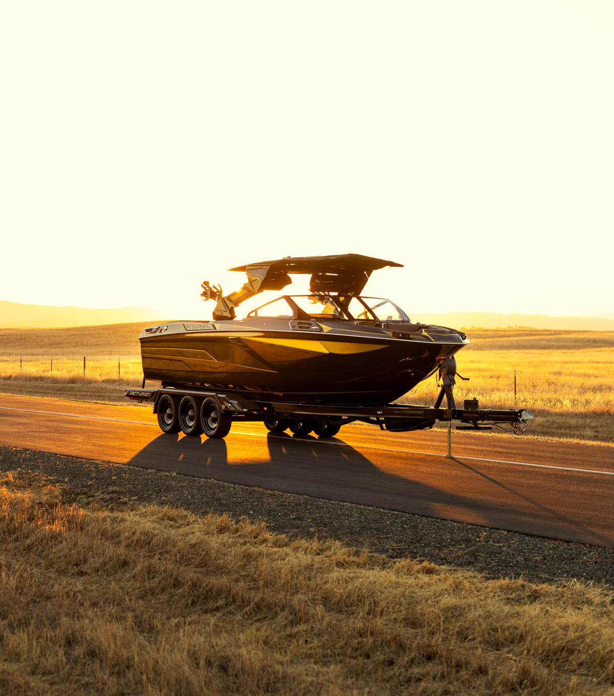 A black Centurion Ri230 wake boat on a trailer is parked on a rural road at sunset, with golden fields and distant hills in the background.