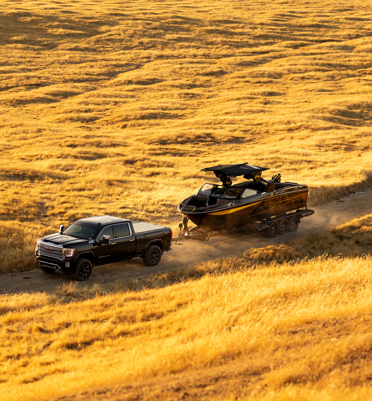 A black pickup truck tows a sleek black and yellow Centurion Ri230 along a dirt path through a dry, golden grass field.