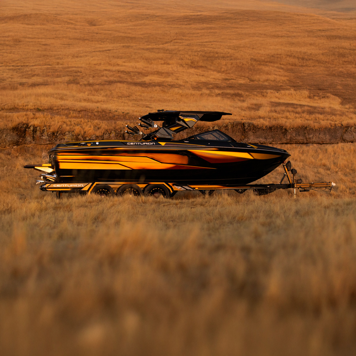 A black and orange Centurion Ri230 Wake Boat on a trailer is parked in a field of tall dry grass with rolling hills in the background.