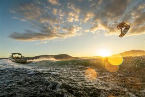 A person wakeboarding is airborne above the water at sunset, with a boat creating waves behind them and hills in the background.