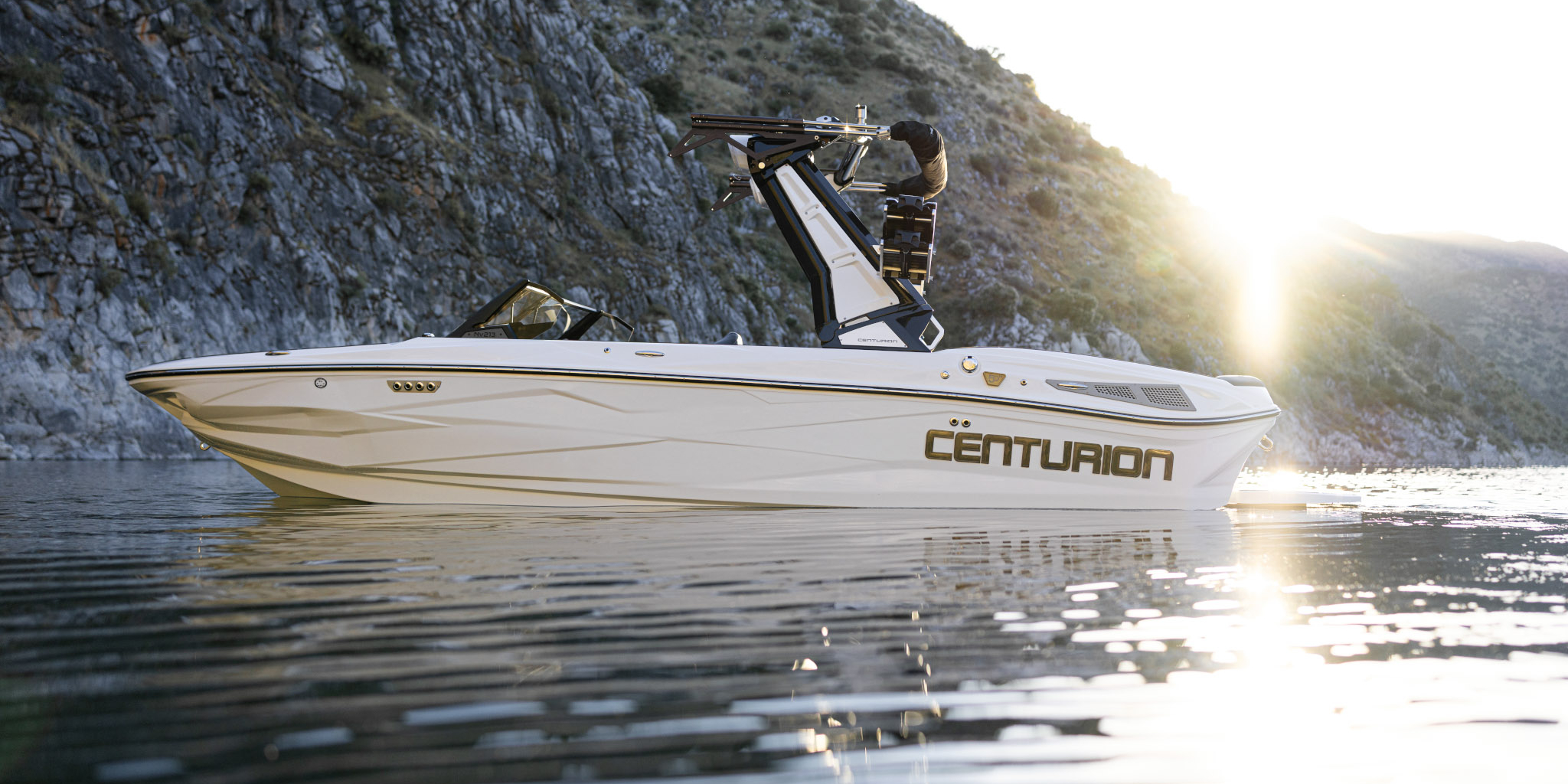A white Centurion speedboat floats on calm water near rocky hills at sunset.
