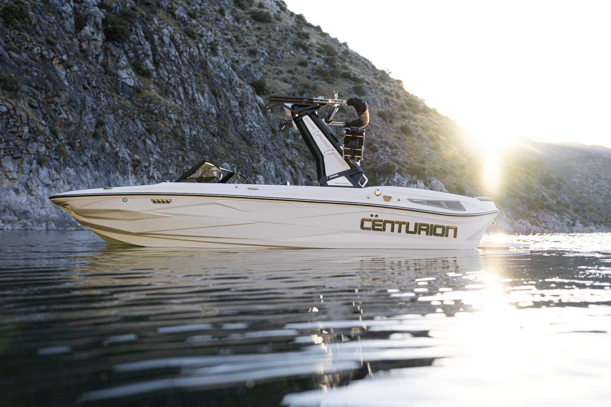 A white Centurion speedboat floats on calm water near rocky hills at sunset.
