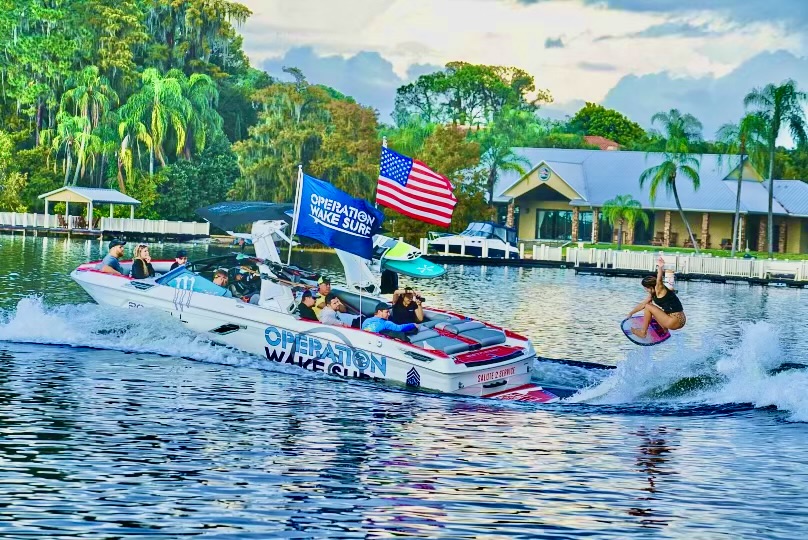 A boat labeled "Operation Wake Surf" pulls a surfer on a lake, with several people on the boat and an American flag displayed, trees and a house in the background.