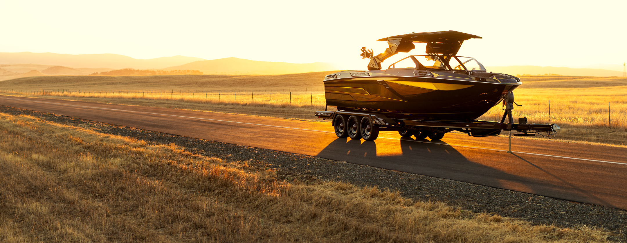 A black Centurion Ri230 Wake Boat on a trailer is parked on a rural road at sunset, surrounded by open fields and distant hills.