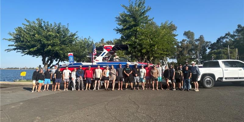 A group of people stands in front of a boat hitched to a white truck at a lakeside parking area, with trees and water visible in the background.