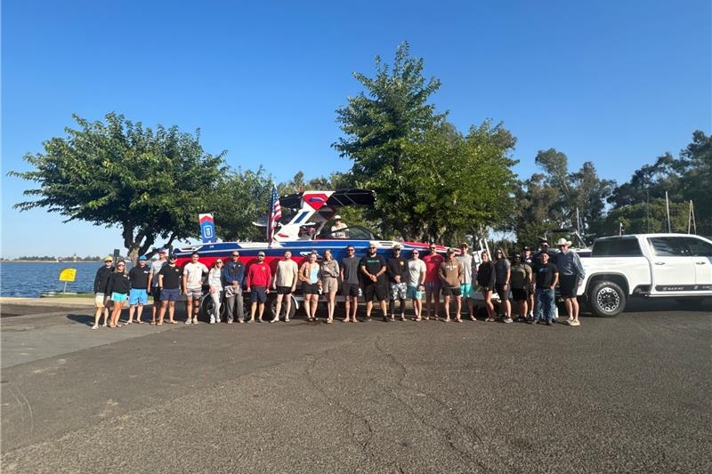 A group of people stands in front of a boat hitched to a white truck at a lakeside parking area, with trees and water visible in the background.