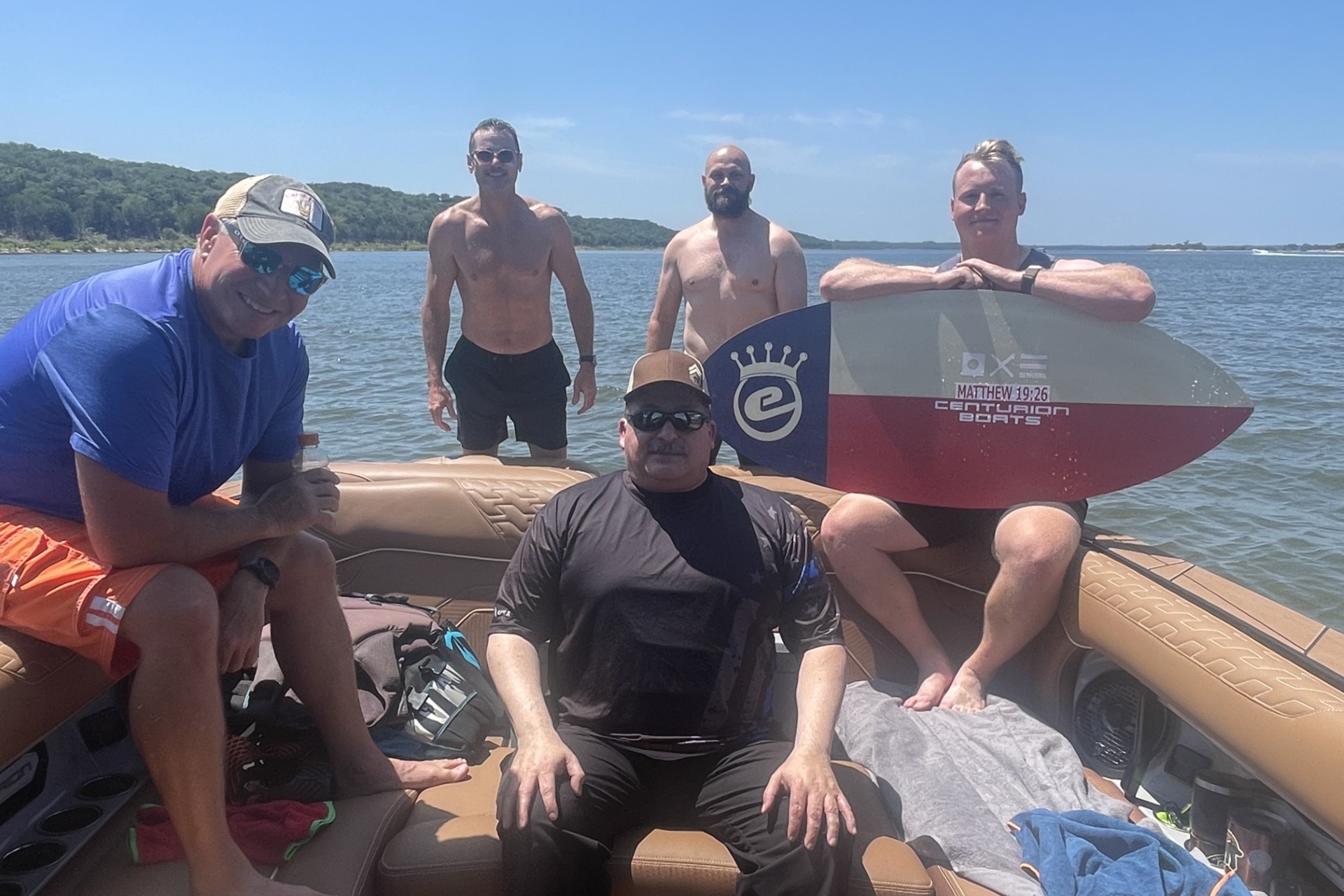 Five men on a boat and in shallow water by a lake, with two standing in the water and three sitting on the boat; one holds a wakesurf board. Trees and shoreline are visible in the background.