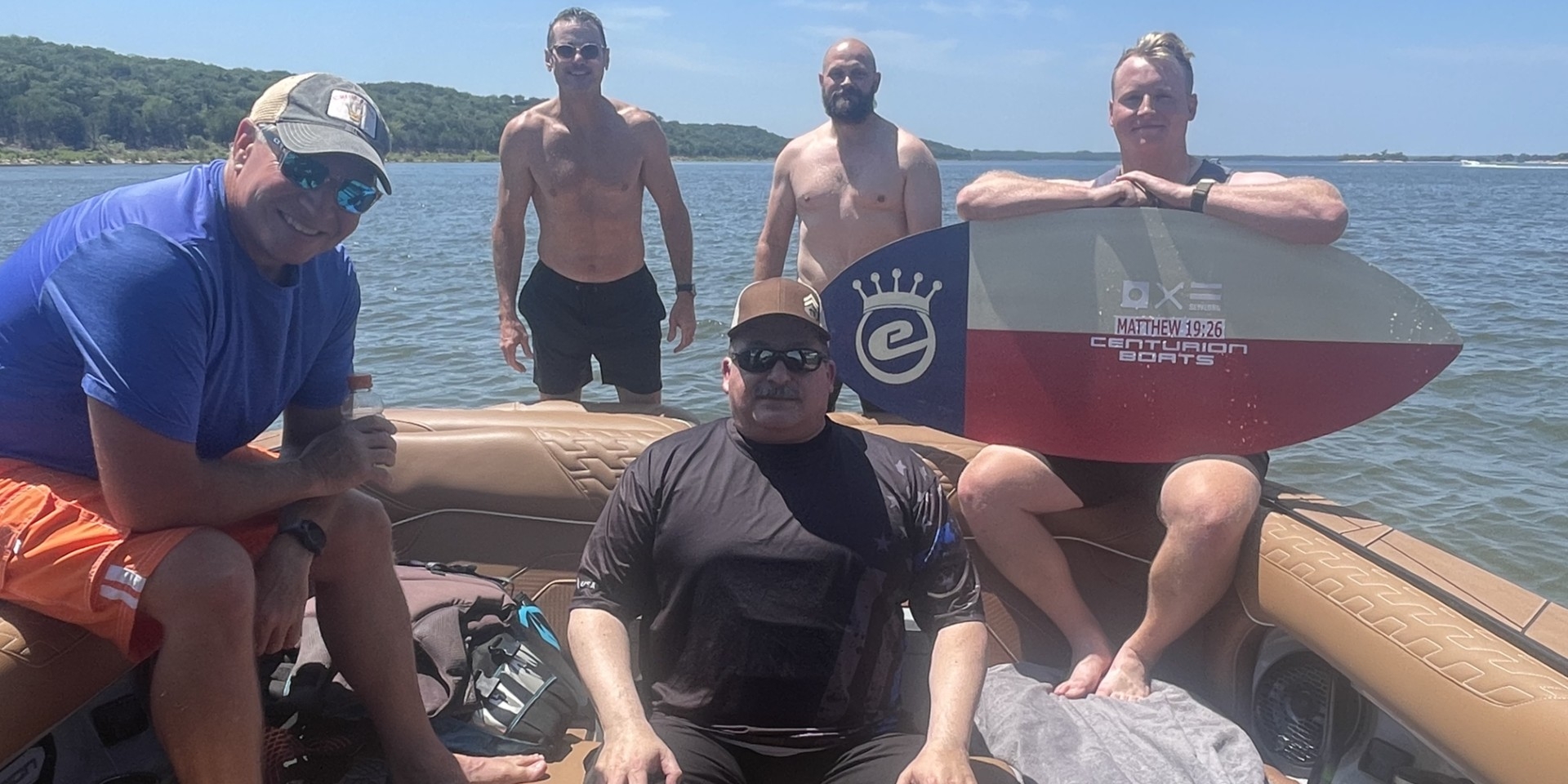 Five men on a boat and in shallow water by a lake, with two standing in the water and three sitting on the boat; one holds a wakesurf board. Trees and shoreline are visible in the background.