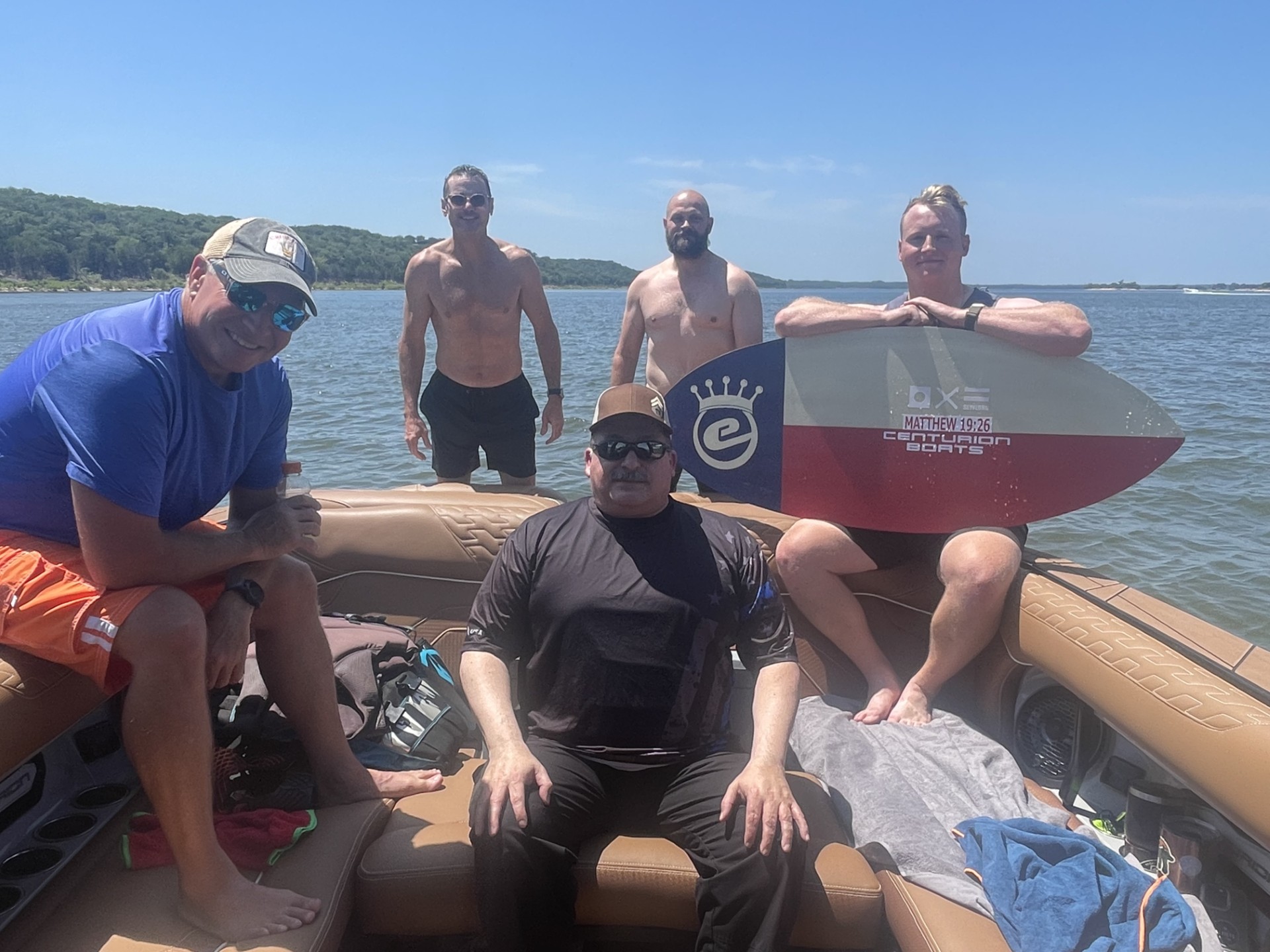 Five men on a boat and in shallow water by a lake, with two standing in the water and three sitting on the boat; one holds a wakesurf board. Trees and shoreline are visible in the background.