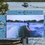 A large outdoor screen displays a wake surfer riding a wave, with event banners for the World Wake Surfing Championship on either side and a boat on the lake in the background.