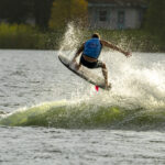 A person wearing a blue vest is wakeboarding on a lake, catching air above the wake with water splashing around them.