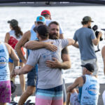 Two men hug near a lakeside event booth, surrounded by people in blue "World Wake Surf" shirts and hats, with water visible in the background.