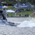 A person in a blue vest is wakesurfing and jumping off the wake behind a Centurion boat, while people watch from the boat and spectators gather on the grassy shore.