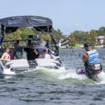A group of people ride in a motorboat pulling a wakeboarder on a sunny day, while another person floats nearby in the water.