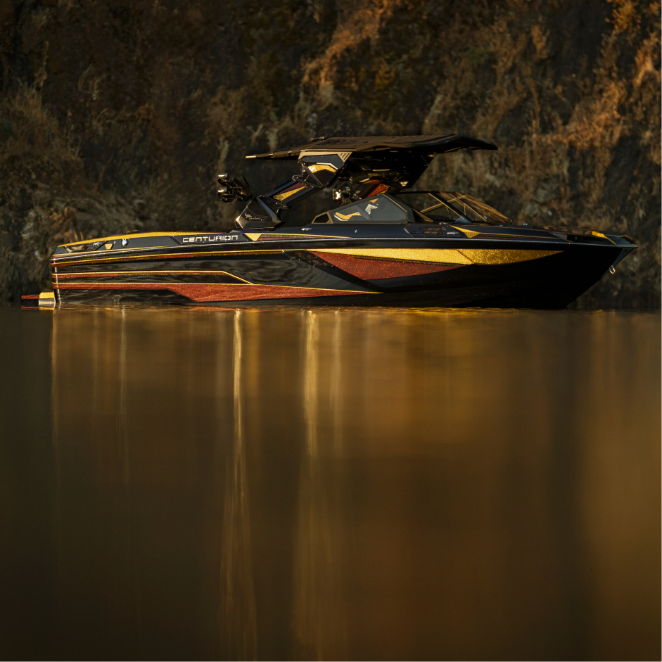A black and gold Centurion boat with the advanced Opti-V Hull floats on calm water near a rocky shoreline with a brown and green backdrop.