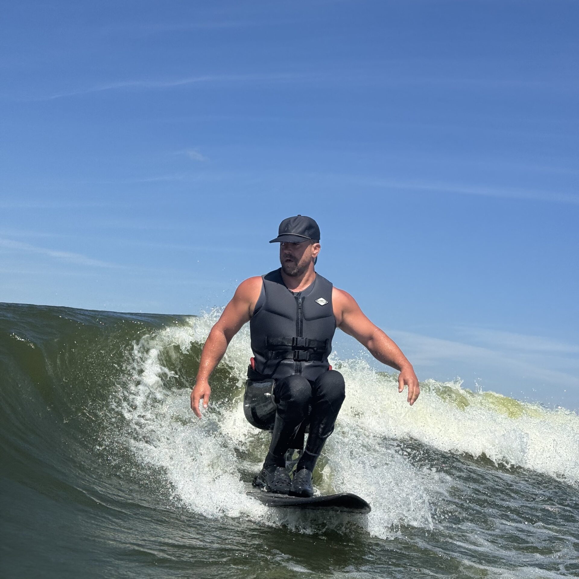 Jordan Koeninger, wearing a life vest and cap, rides a wave on a surfboard under a clear blue sky.