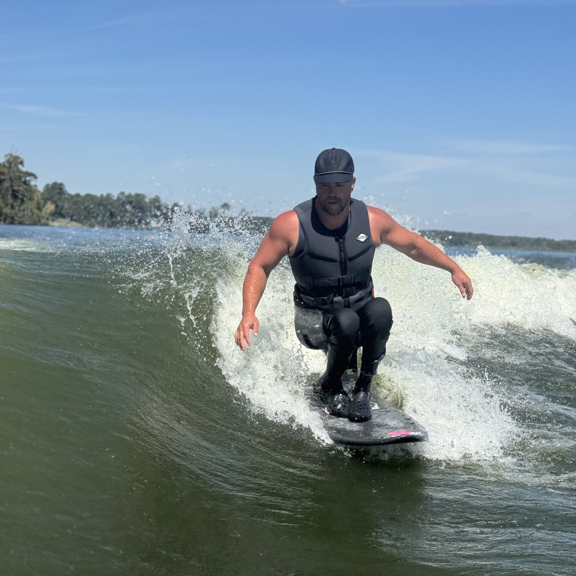 A man wearing a life vest and cap is wakesurfing on a wave under a sunny blue sky.