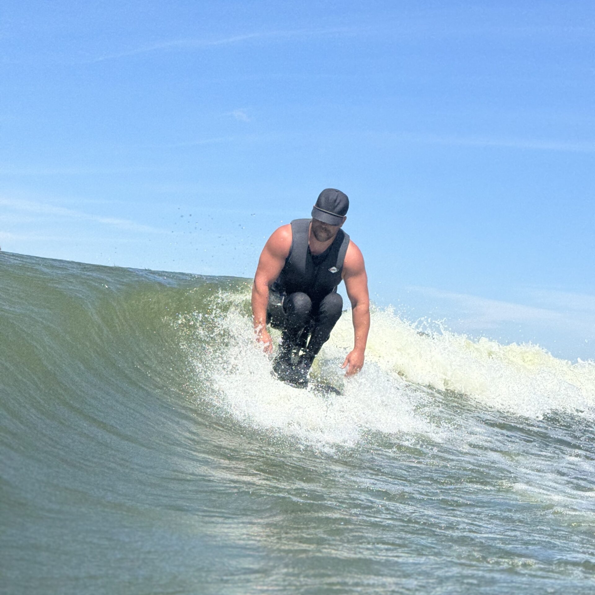 Jordan Koeninger, wearing a cap and sleeveless wetsuit, is surfing on a wave under a clear blue sky.