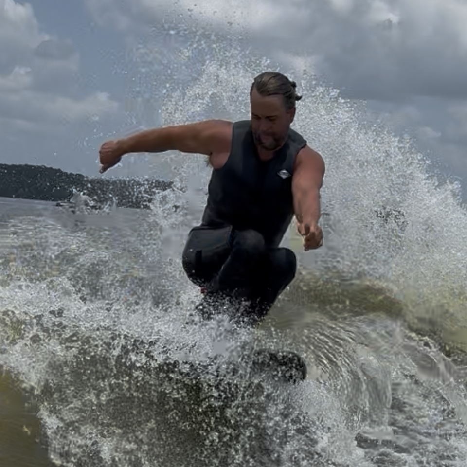 A person wearing a life vest skims over the surface of the water, creating a large splash against a backdrop of cloudy sky and distant trees.