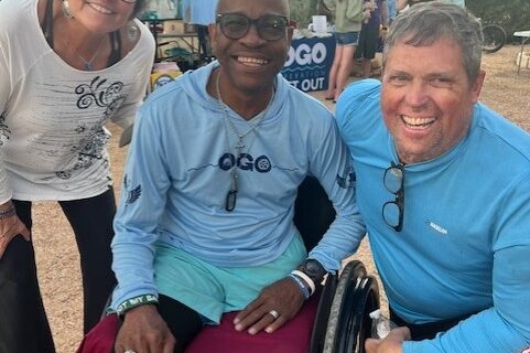 Three adults smile at an outdoor event booth; one is seated in a wheelchair, and two are standing on either side. They all wear casual, sporty clothing.