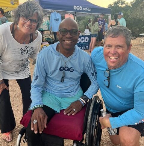Three adults smile at an outdoor event booth; one is seated in a wheelchair, and two are standing on either side. They all wear casual, sporty clothing.
