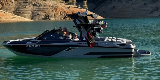 A black and gray speedboat with two people onboard floats on a green lake near a rocky, tree-lined shore.