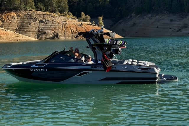 A black and gray speedboat with two people onboard floats on a green lake near a rocky, tree-lined shore.