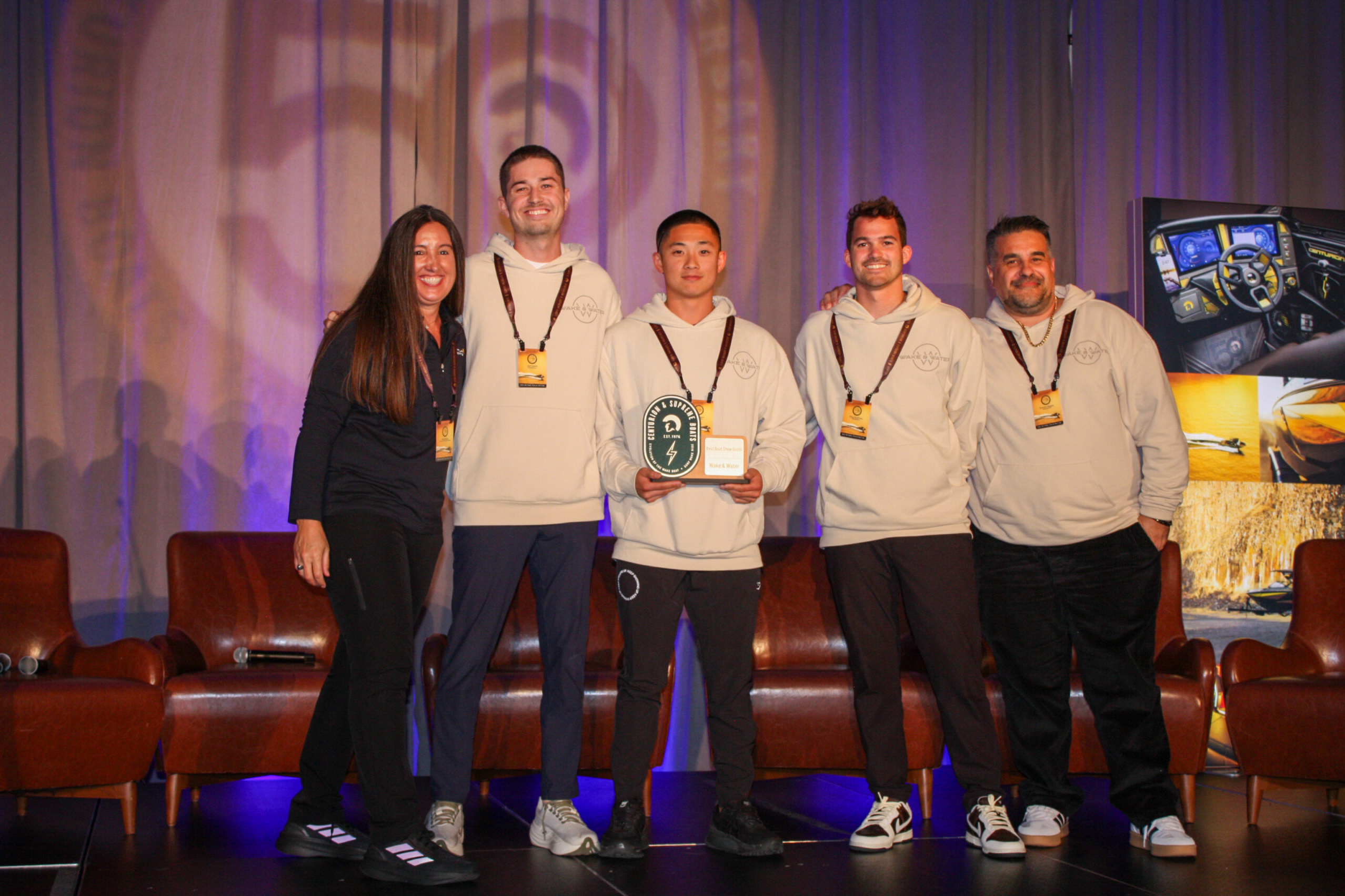 Five people stand on a stage, with one person in the center holding a plaque and small box. All are smiling and wearing event badges; three wear matching light-colored hoodies.