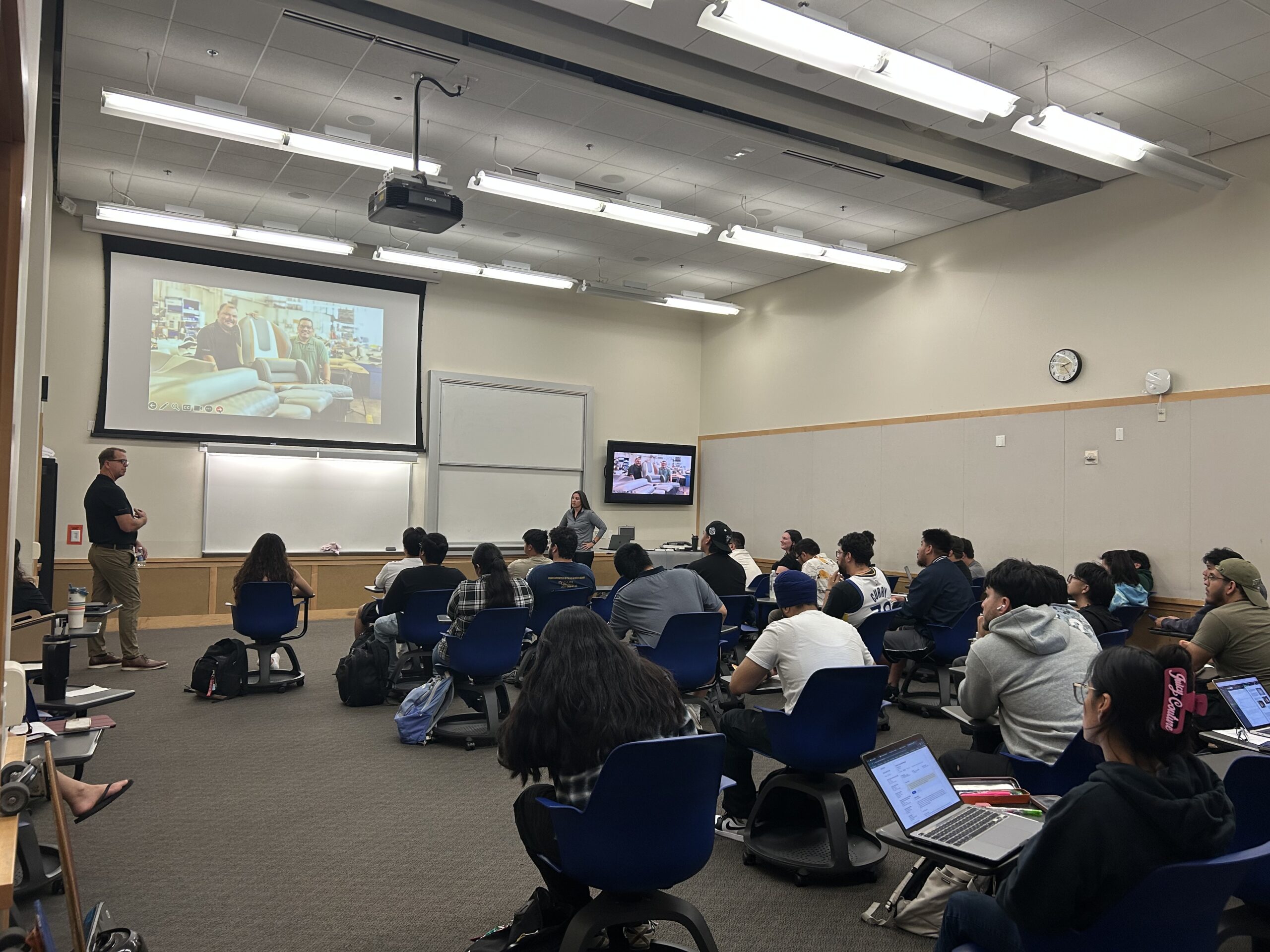 A classroom with students seated at desks facing a lecturer and presentation screen; some students use laptops while listening.