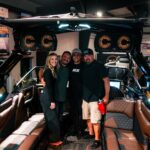 Four people stand and pose together inside a luxury motorboat displayed indoors, surrounded by leather seating and illuminated speakers.