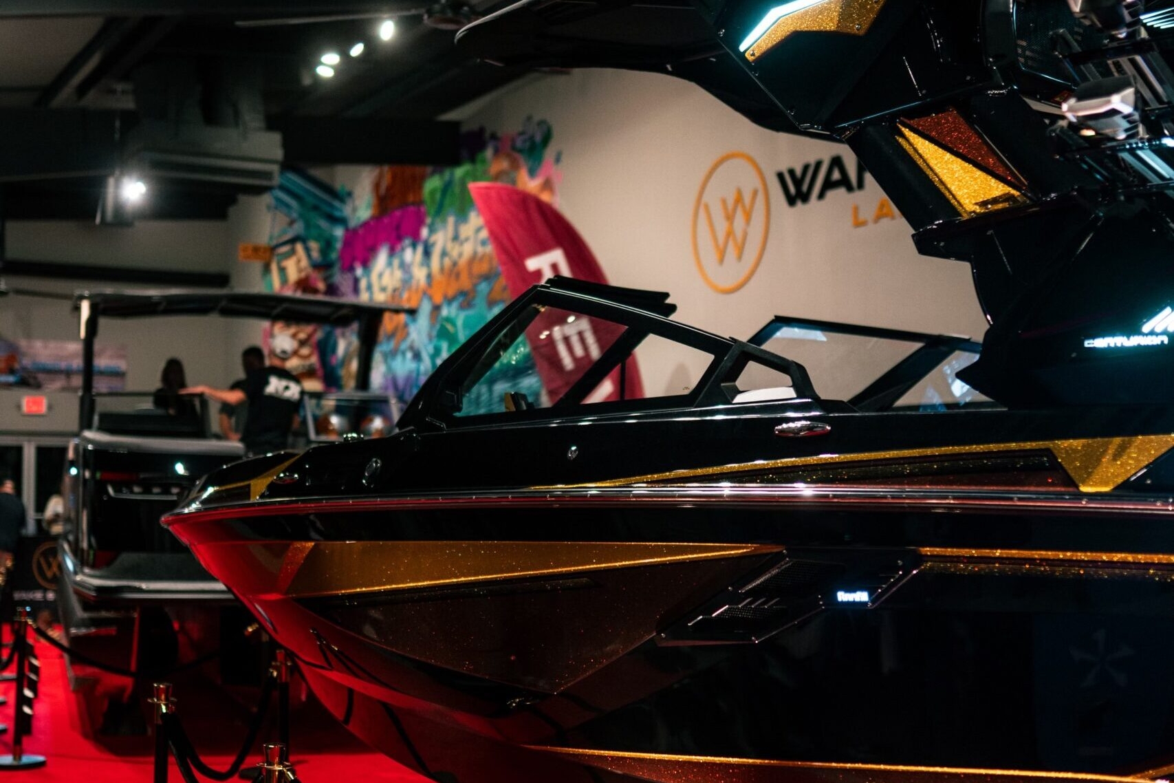 A black and gold speedboat is displayed indoors on a red carpet, with ropes and stanchions surrounding it and bright lights overhead.