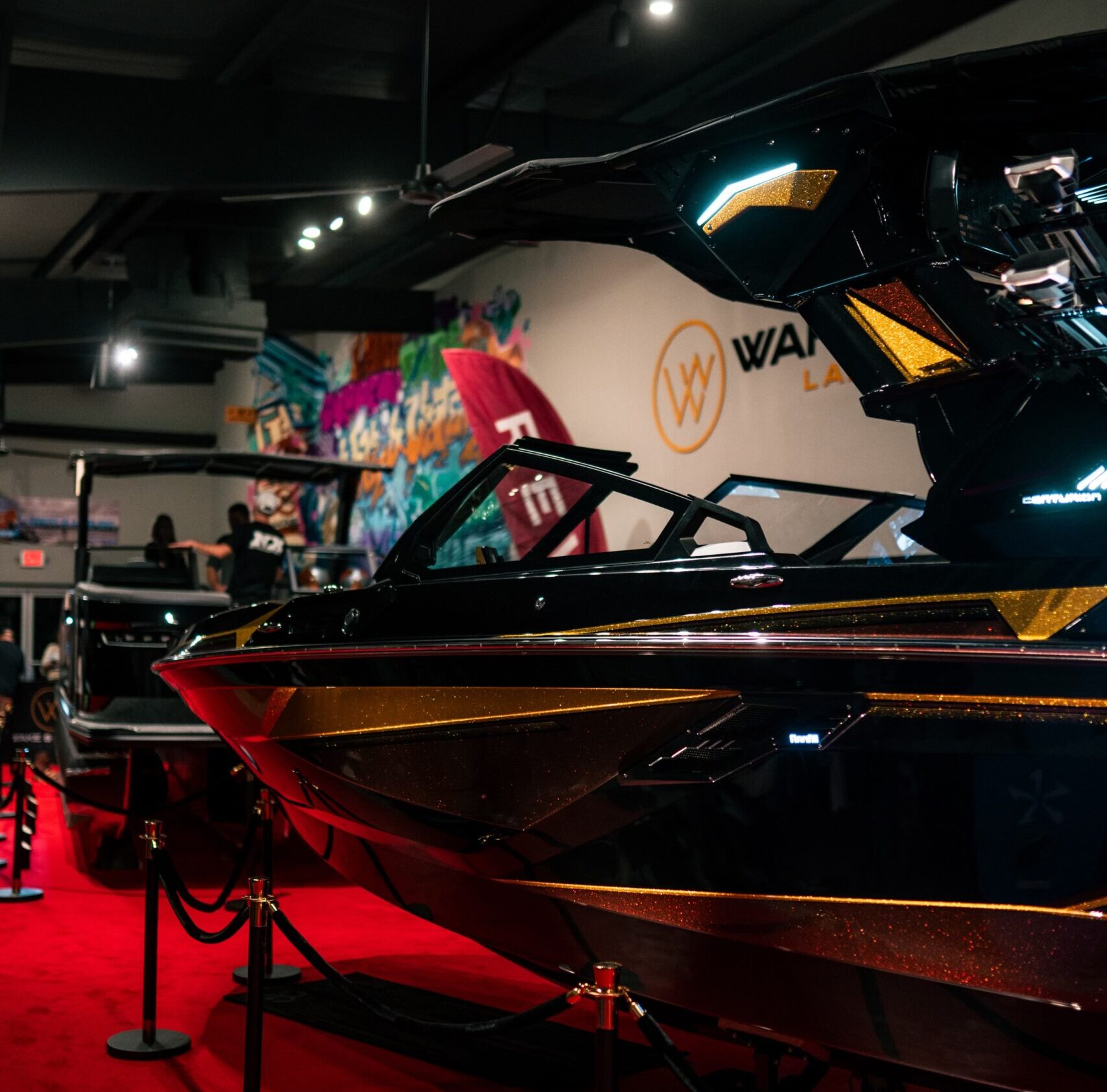A black and gold speedboat is displayed indoors on a red carpet, with ropes and stanchions surrounding it and bright lights overhead.