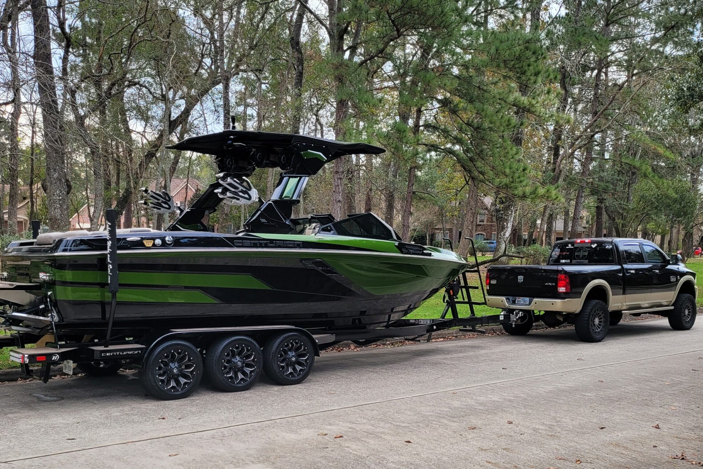A black pickup truck is towing a large black and green speedboat on a trailer along a residential street lined with trees.