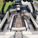 Interior view of a modern boat with beige and gray cushioned seating, wooden floor panels, and audio speakers, parked near a black pickup truck.