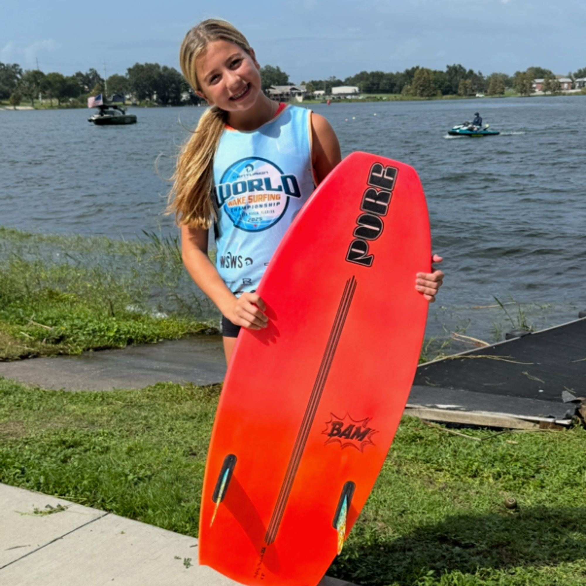 Young girl, Clair Hurta, stands by a lakeshore holding a bright orange wakesurf board, wearing a sleeveless competition shirt, with watercraft and trees visible in the background.