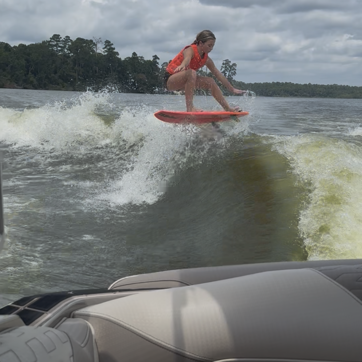 Tripp Allen, in a red life jacket, wakesurfs on a lake, riding a wave behind a boat beneath a cloudy sky.
