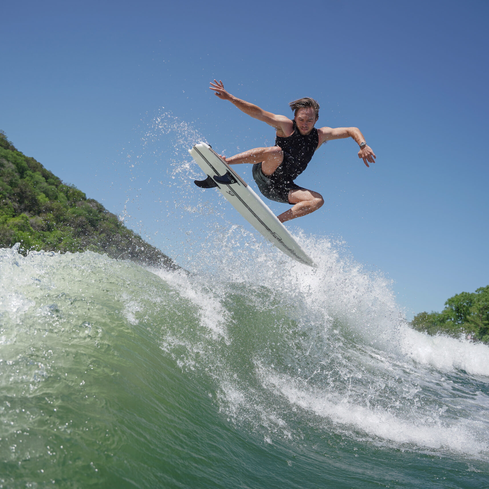Trevor Miller, in a black wetsuit, jumps with a surfboard above a wave near a green, tree-lined shore under a clear blue sky.