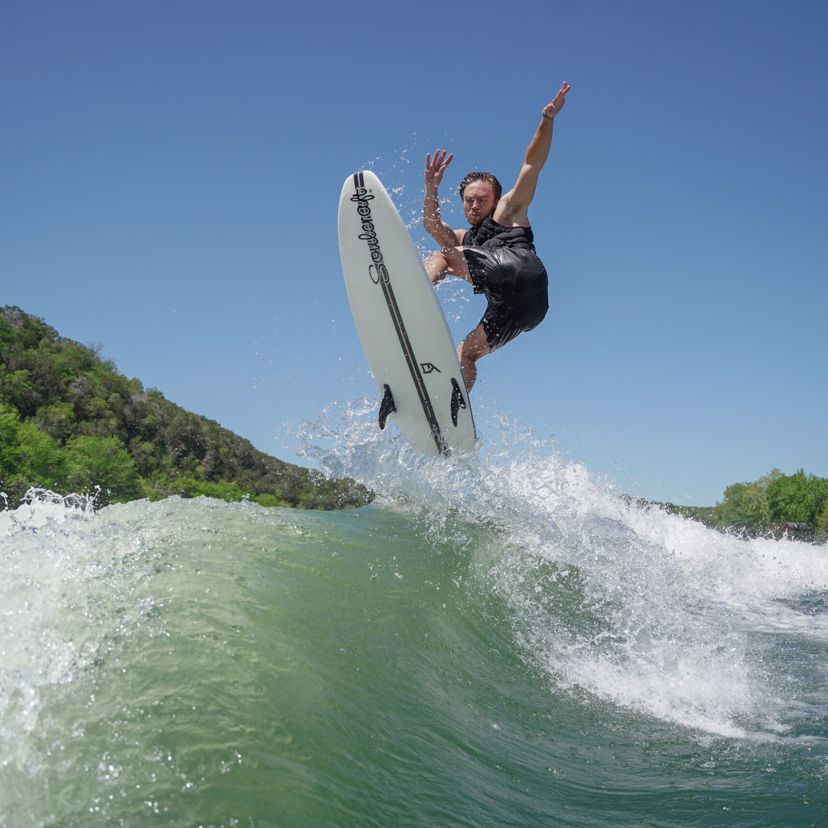 Trevor Miller, in a wetsuit, is airborne on a surfboard above a wave, surrounded by green trees and a clear blue sky.