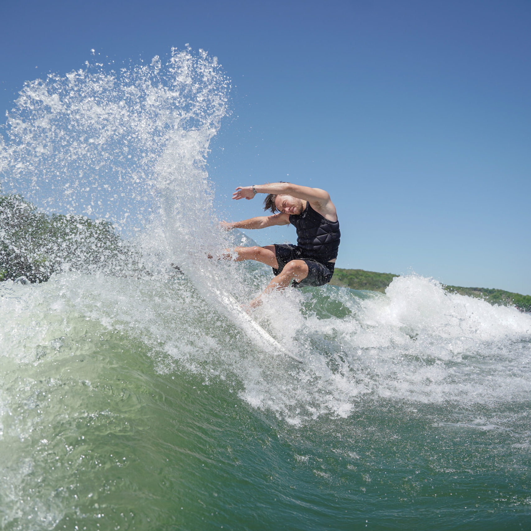 Trevor Miller, in a black life jacket, surfs on a wave, kicking up water under a sunny blue sky with trees in the background.