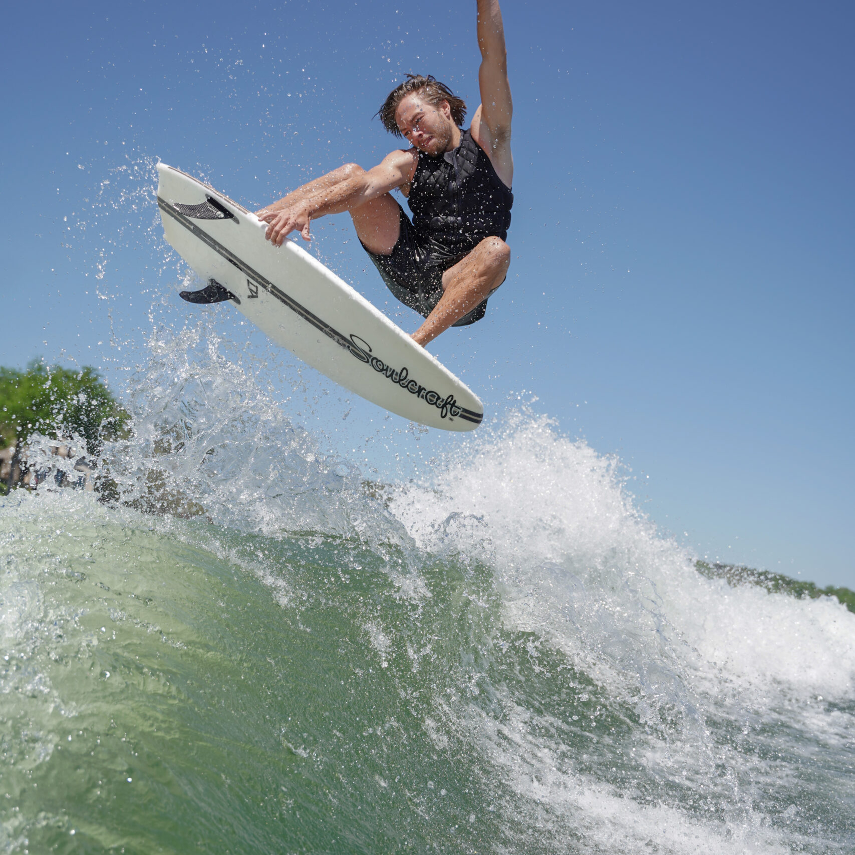 Trevor Miller performs an aerial trick on a surfboard above a wave, wearing a black tank top and shorts, with one hand making a peace sign against the clear blue sky.