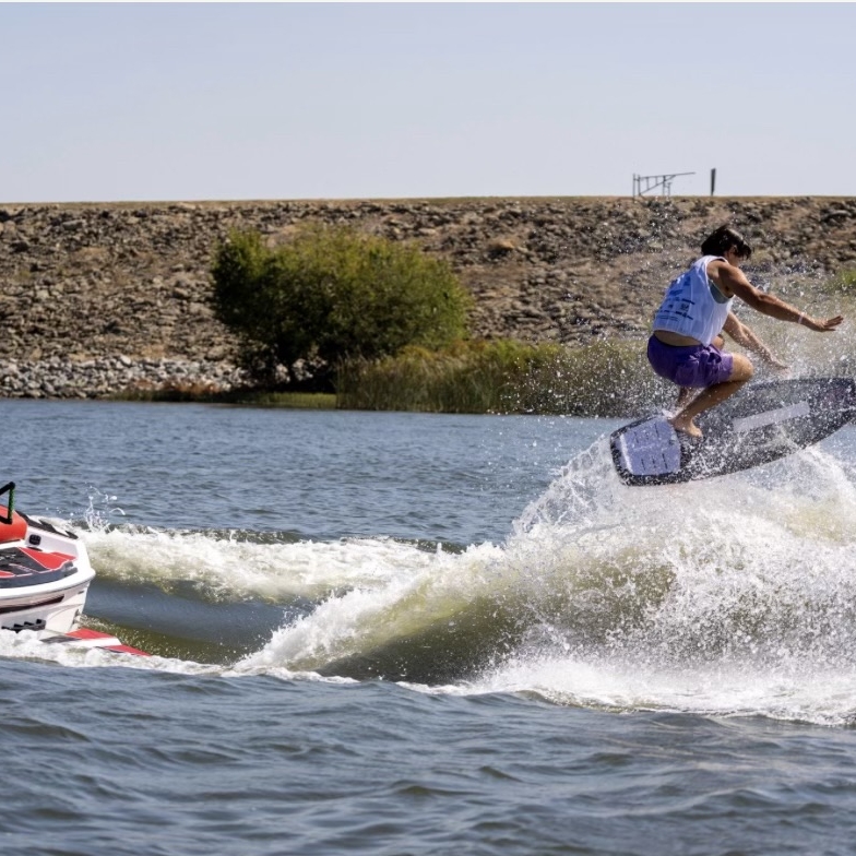 Jordan Koeninger, in a life vest, wakeboards behind a red and white boat, performing an impressive jump over a wave on a sunny day.