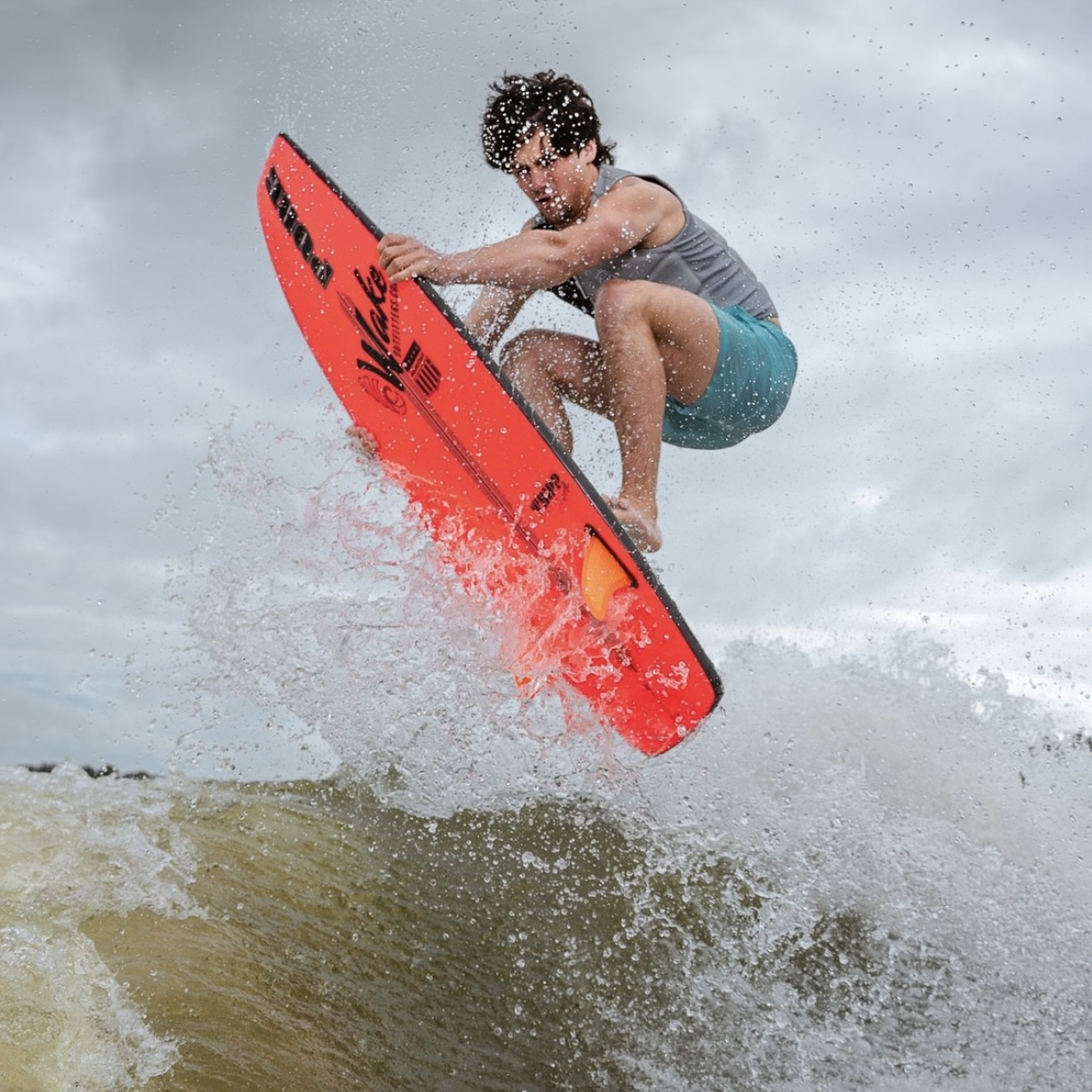 Jordan Koeninger, in a gray tank top and teal shorts, performs a jump on a bright red surfboard above a wave, water splashing around him under a cloudy sky.